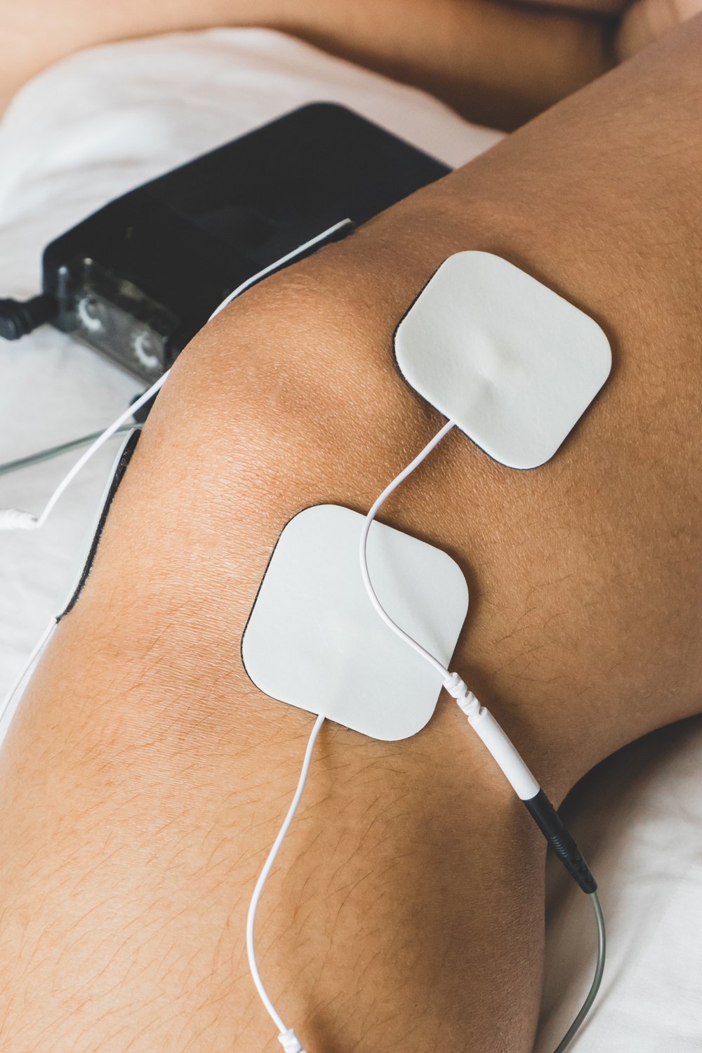 Close-up of electrical muscle stimulation (EMS/TENS) pads attached to a person's knee, connected by wires to a small black stimulator unit.